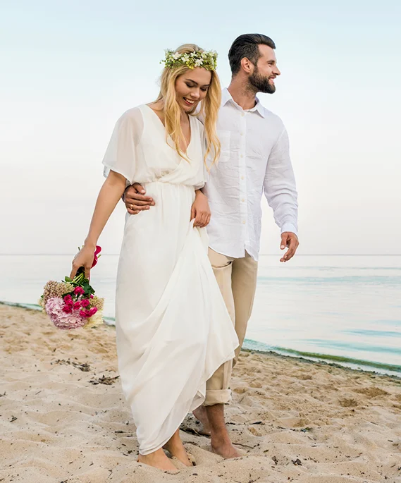 A happy couple walks barefoot along a sandy beach by the ocean. The woman wears a flowy white dress and a flower crown while holding a bouquet of flowers, and the man walks beside her with his arm around her waist.