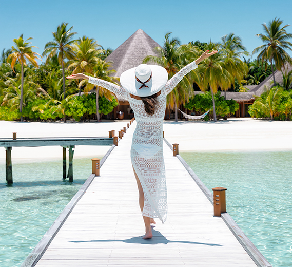 A woman from behind with arms outstretched, wearing a white sunhat and sheer dress, walks down a wooden pier toward a tropical resort beach.