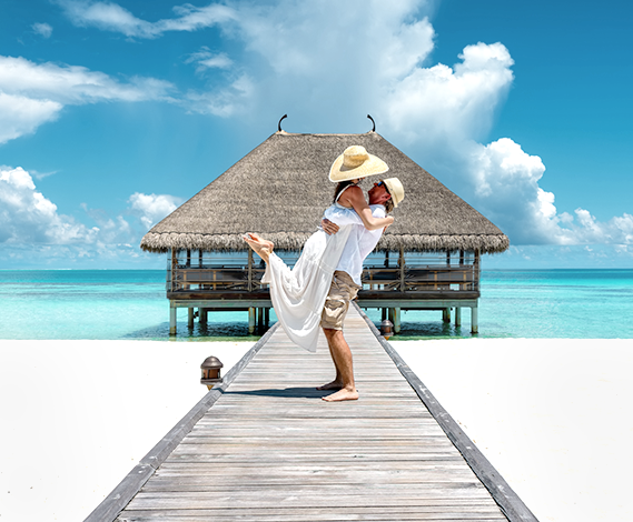 A romantic couple embraces on a wooden pier leading to a thatched-roof overwater bungalow, surrounded by white sand and turquoise tropical water under a blue sky.